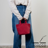 一位身穿牛仔褲與淺色長袖上衣的模特兒，手提一個紅色的三河木綿刺子織圓底水桶包。