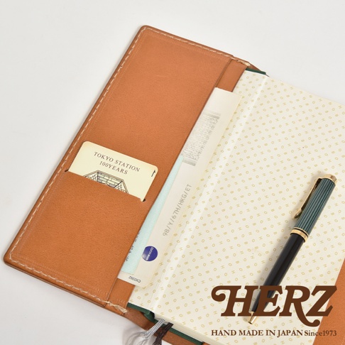 職人手作皮革旅行記事本書套，採用棕色皮革製作，內含卡片夾層，可放置卡片、機票等物品。書套內頁印有規則的圖案，搭配一支綠色筆，展現簡約質感。