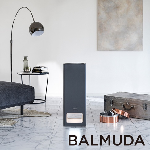 The Pure 空氣清淨機，機身為深灰色，造型為長方體，底部有白色發光面板，置於淺色大理石紋地板上，旁邊有復古風格的金屬行李箱和銅色飾品。