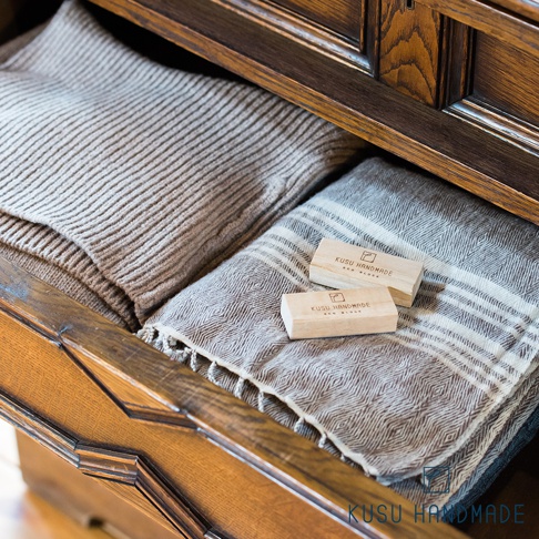 兩個淺木色樟楠木條，印有「KUSU HANDMADE ECO BLOCK」字樣，放置在深棕色木質抽屜內，抽屜內有兩件淺棕色和灰白條紋的織物。