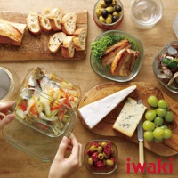 透明玻璃微波保鮮盒內盛有魚和蔬菜，盒蓋被拿起，旁邊擺放著麵包、烤肉、起司、葡萄和橄欖。