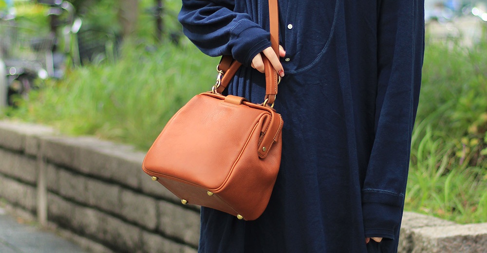 棕褐色皮革方形口金托特包，附可拆卸肩帶，包身有金色金屬底釘和金屬扣環，由身著深藍色服飾的人士斜肩背著。
