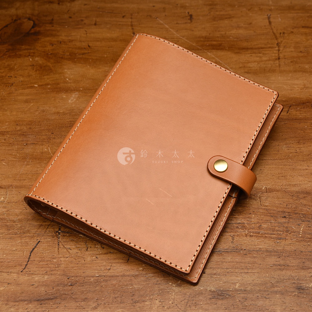 職人手作皮革A5手帳/記事本書套，淺棕色牛皮材質，邊緣有縫線裝飾，前方有皮質繫繩與金色金屬釦固定。