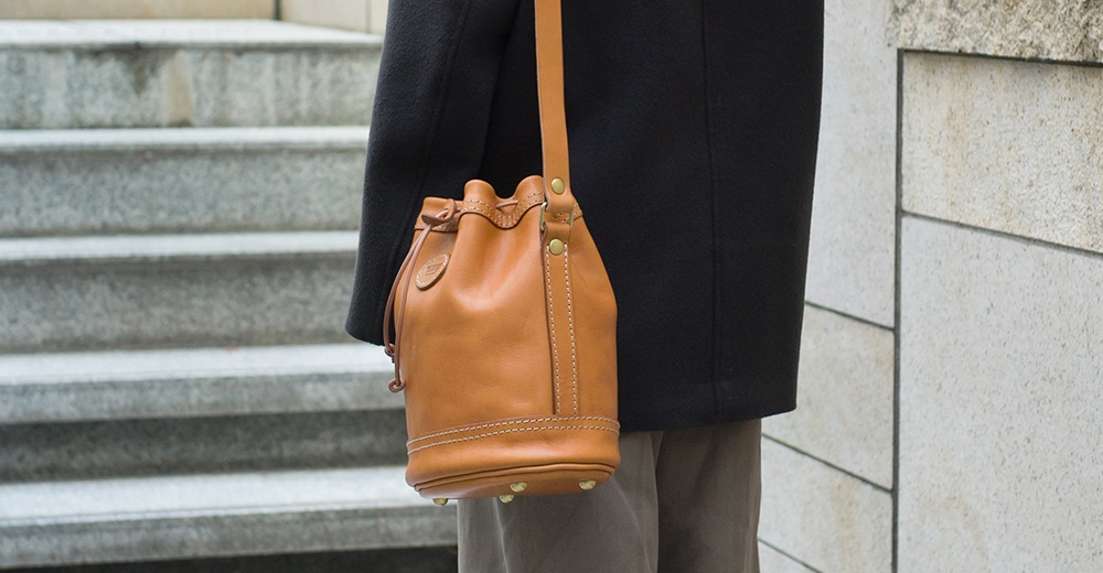 棕色皮革束口單肩包，配有可調節背帶、束繩開口及金色金屬腳釘，包身有縫線細節。