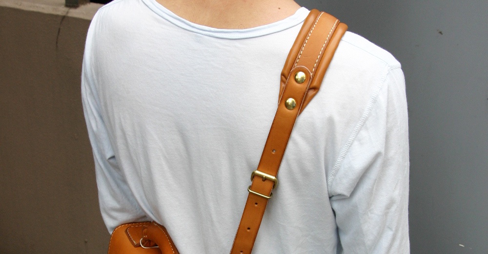 棕褐色皮革加厚減壓肩背帶，搭配金色釦環及鉚釘，肩帶上有多段調整孔，縫線清晰。