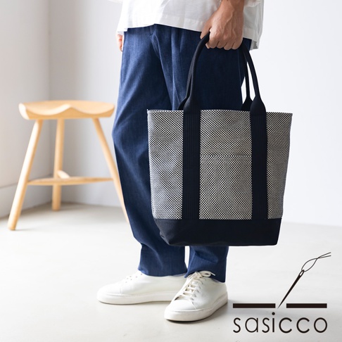 一款黑色與白色格紋刺子織托特包，配有黑色提把和底部，由三河木綿製成，由一人手持。