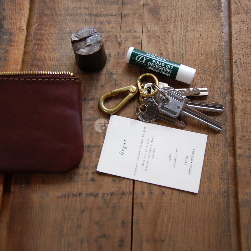 酒紅色皮革拉鍊收納袋，搭配黃銅色登山扣與鑰匙串、潤唇膏及一枚圓形金屬壓印模，一同擺放在木紋桌面上。