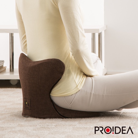 深棕色馬鞍型正姿座墊，材質為織物，具有支撐臀部與背部的設計，一位身穿米黃色上衣與淺灰色褲子的女士正坐於座墊上。