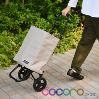 米色帆布材質的2WAY手提收納折疊推車，配有黑色金屬骨架和輪子，正在由一人拉行通過鋪砌的戶外路徑。