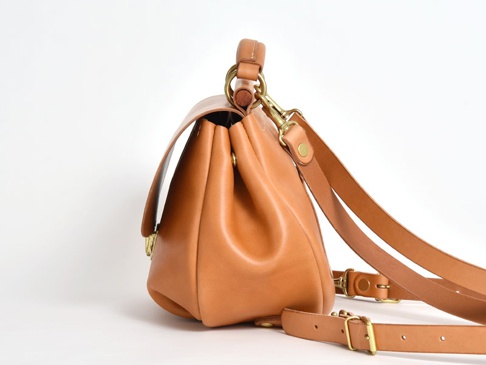 職人手作皮革屋瓦風迷你後背包，採用天然棕色皮革製成，帶有復古金色金屬扣環和可調節肩帶。包身側面呈現圓潤的線條，頂部提把設計。