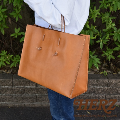 職人手作皮革提袋 T-5，採用棕色皮革製成，有兩個細長皮革提把，包身正面有兩個以皮革繩綁成的裝飾結。