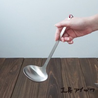 一只手正握著一個不鏽鋼湯勺，湯勺勺頭為圓弧形，勺柄細長，整體為銀白色金屬光澤。