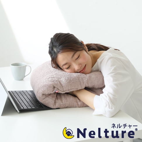 一位女子趴在淡粉色毛巾材質的甜甜圈形狀枕頭上休息，枕頭放在桌面上，旁邊有筆記型電腦和杯子。