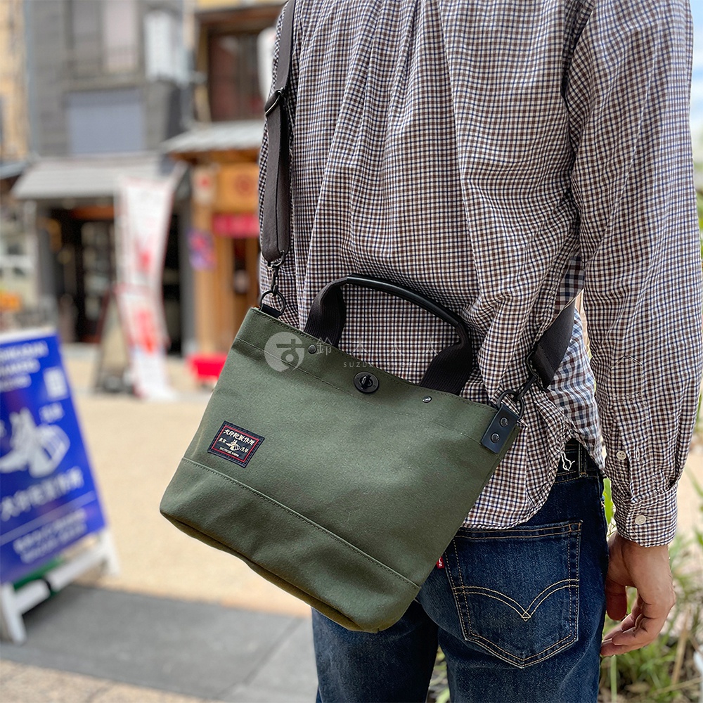 深綠色帆布材質的兩用托特包，具有手提帶及可拆卸肩帶，包款正面有品牌標誌布章。