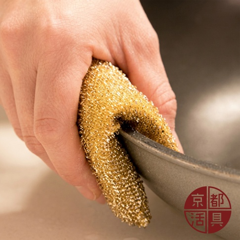 一隻手握著金色的長條狀清潔菜瓜布，正對著一個灰色鍋具邊緣進行刷洗，底部右下角印有紅色的京都活具品牌標誌。