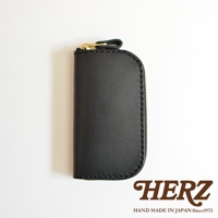 黑色皮革橢圓形鑰匙包，頂部有金色拉鍊和皮革拉環，包體縫線精緻，右下角印有HERZ字樣及HAND MADE IN JAPAN Since1973字樣