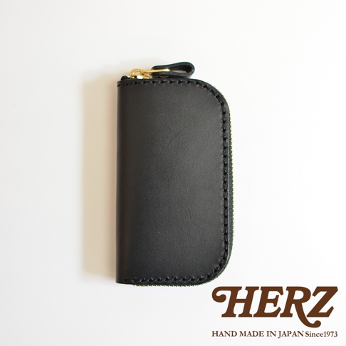 黑色皮革橢圓形鑰匙包，頂部有金色拉鍊和皮革拉環，包體縫線精緻，右下角印有HERZ字樣及HAND MADE IN JAPAN Since1973字樣