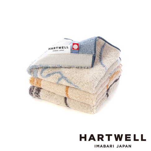 三條摺疊整齊的 Hartwell 今治毛巾手帕堆疊在一起，材質呈現柔軟的毛圈質感，以米色為底色，綴有藍色、黃色與灰色的線條圖紋，毛巾邊緣縫有印著 Hartwell Imabari Japan 字樣及紅白色標籤的品牌識別標記。