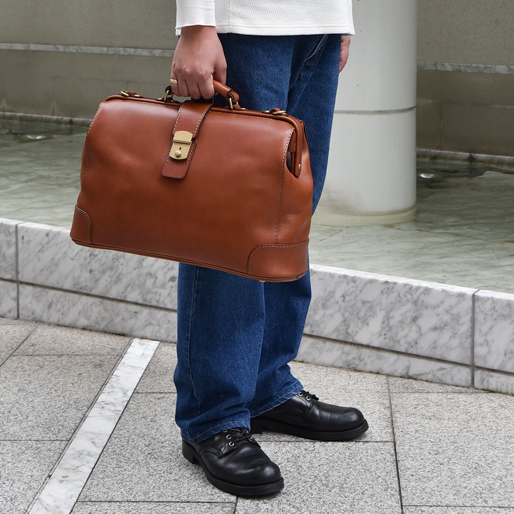 職人手作皮革商務旅行兩用公事包，呈經典醫生包造型，光滑的棕褐色牛皮材質，配有金色金屬鎖扣和提把。