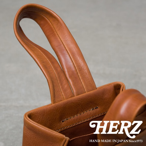 職人手作的棕色皮革單柄手提包，包身呈現自然皮革紋理，提把部分設計簡潔，包口邊緣有縫線細節，展現皮革質感。