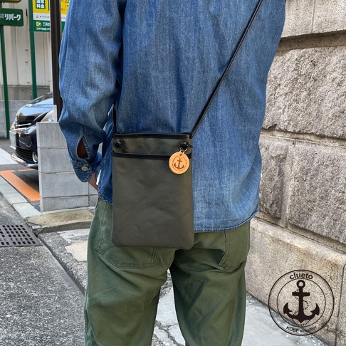 一款墨綠色帆布皮革散步包，搭配黑色皮革飾邊與繫繩，包包上掛有船錨圖案的木製吊牌，由一位身穿牛仔襯衫、綠色長褲的男性斜背展示。
