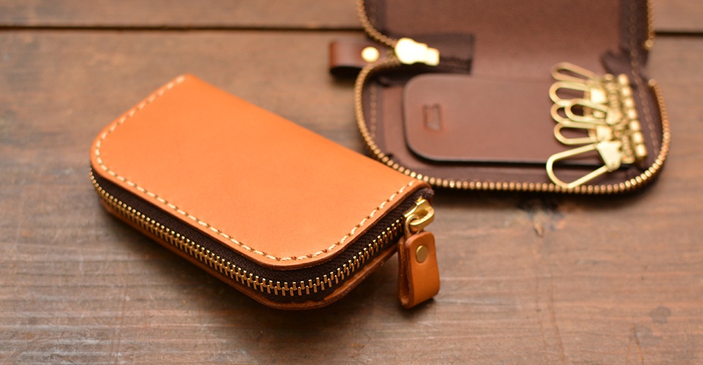 職人手作的橘棕色皮革橢圓形拉鍊鑰匙包，拉鍊處有著金色五金和皮革拉環，包內有六個鑰匙鉤和一個皮革卡片夾，背景為木紋桌面。
