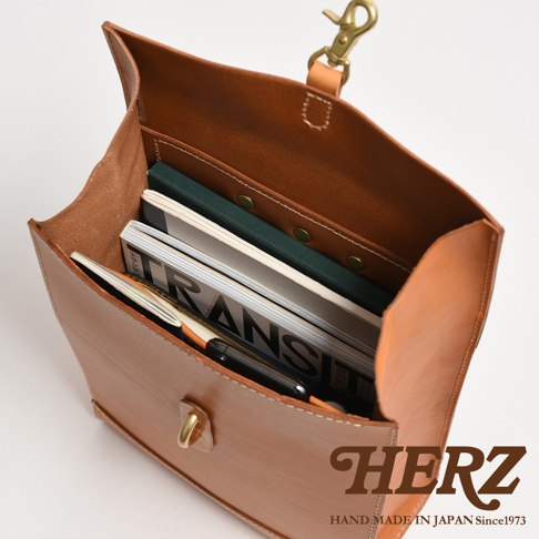 淺棕色皮革紙袋風後背包，內部裝有雜誌、筆和綠色筆記本。包款可見黃銅色扣環和鉤子，以及品牌Logo。