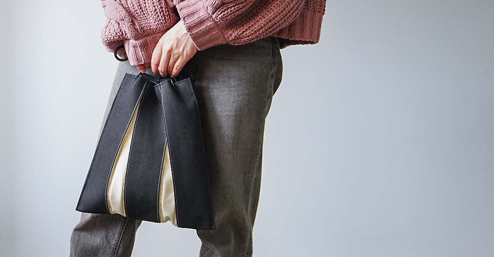 一位穿著粉色針織毛衣與灰色牛仔褲的女性，手持京都KOSHO品牌的ougi帆布信玄袋，這款手袋採用黑白雙色拼接設計，具有獨特的扇形摺疊結構與簡約俐落的風格。