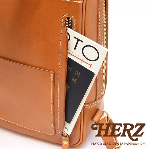 職人手作皮革長方型多口袋後背包，展示部分為淺棕色皮革，側邊拉鍊口袋內插有白色封面、黑色內頁的物品。