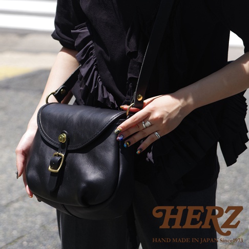 一位女士肩背著一個日本 HERZ 品牌的黑色手工皮革休閒旅行馬鞍包，包款設計簡約，配有金色釦環和錶帶，包包的局部細節與女士的手部裝飾清晰可見。