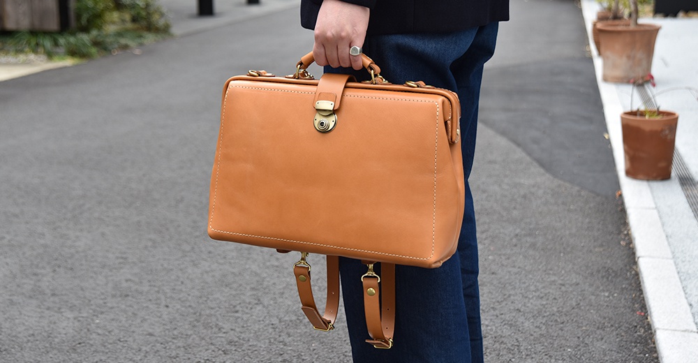 職人手作皮革軟式杜勒斯三用背包 BJ-16，採用淺棕色皮革，設有金色旋轉鎖扣和手提把，可變換為背包模式，肩帶為皮革材質。