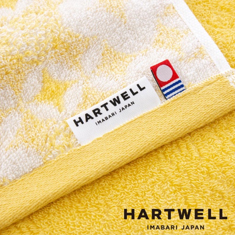 黃色和白色相間的毛巾，帶有傳統日式紋樣，材質為柔軟的棉質。毛巾上有「HARTWELL IMABARI JAPAN」的品牌標籤，以及一個紅色、白色和藍色的裝飾標識。
