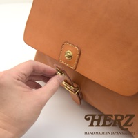 職人手作皮革屋瓦風後背包 R-15，採用天然皮革材質，呈現經典駝色。特寫聚焦於包款的掀蓋設計，展示了精緻的黃銅旋轉鎖扣與皮革繫帶，以及手部正在操作鎖扣的細節。