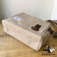 米色純棉帆布製成的橫式側背包，配有咖啡色皮革裝飾和金屬扣環，包蓋上印有品牌標籤。