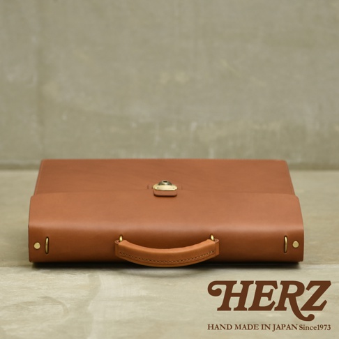 職人手作皮革Organ兩用商務公事包 G-93，採用鞣製皮革製成，呈現溫潤的棕褐色。包款設計簡潔，配有金色金屬鎖扣和手提把，側面有鉚釘裝飾。