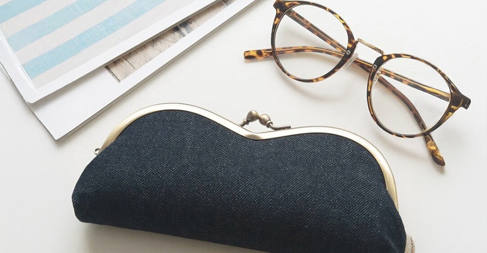 一個帶有虎紋鏡框的眼鏡和一個深藍色牛仔布料的零錢包，擺放在白色的桌面上，旁邊還有一疊有藍色條紋的紙張。