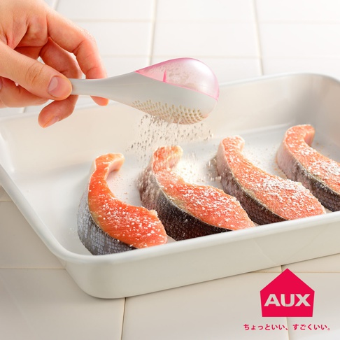 手持白色帶粉紅色透明蓋的leye 料理專用撒粉器，正將細粉狀調味料均勻撒在烤盤中的四塊鮭魚排上。撒粉器底部有細密孔洞，粉末從中落下。