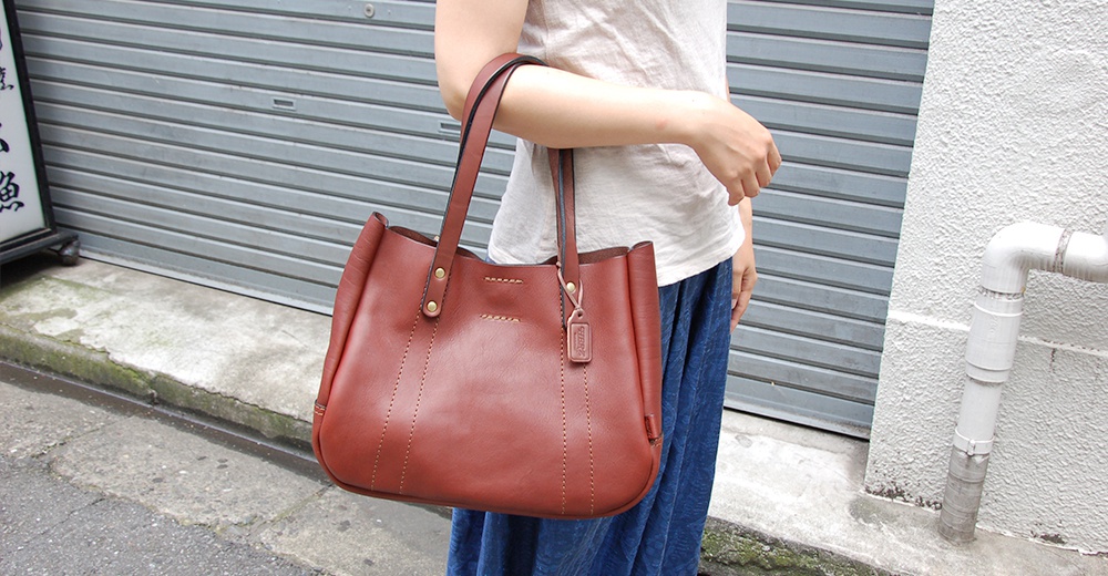 一名女子肩背一個棕色皮革托特包，包包有兩條肩帶，正面有縫線裝飾，右側掛有一個皮革小吊牌。