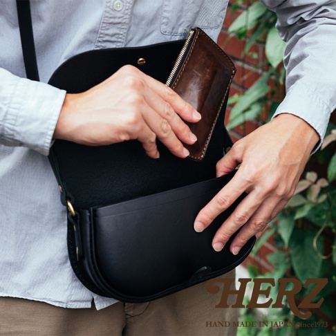 職人手作皮革迷你馬鞍包，黑色，皮革材質。包包為半月形，有可調節的肩帶和金屬扣環。一名男子正在將一個棕色皮革長夾放入包中。