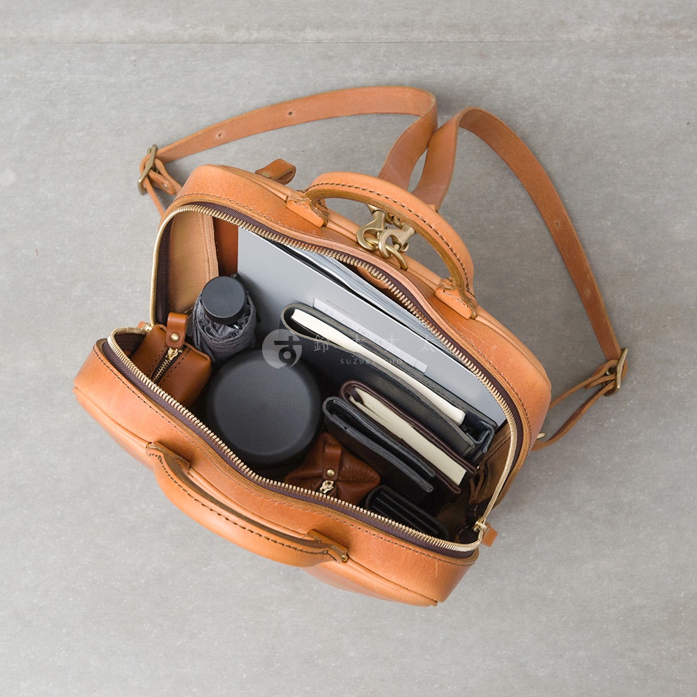 職人手作皮革Organ商務後背包，打開的內部展示了筆記本、文件夾、雨傘、圓形收納盒和錢包等物品，背包採用棕色皮革製成，配有黃銅色五金配件。