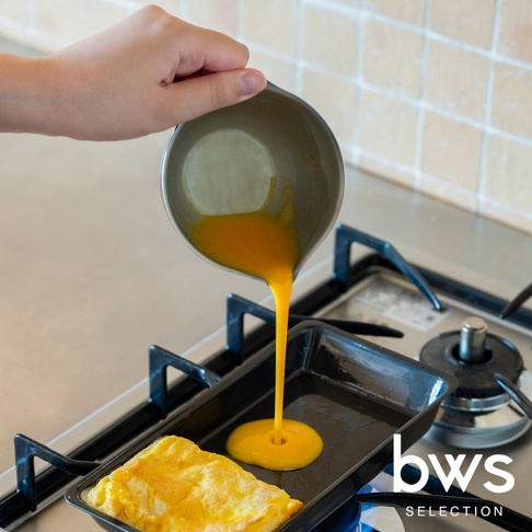 一隻手拿著 bws SELECTION 不鏽鋼料理備料盆，將盆中攪拌好的蛋液倒入正在加熱的長方形平底鍋中，平底鍋內已有一塊煎好的蛋捲，背景為廚房爐台與磁磚牆面。