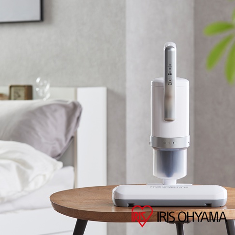 白色與銀色搭配的直立式除塵蟎機，具有銀色提把和透明集塵筒，底部為銀灰色扁平吸頭，放置於木紋圓桌上。