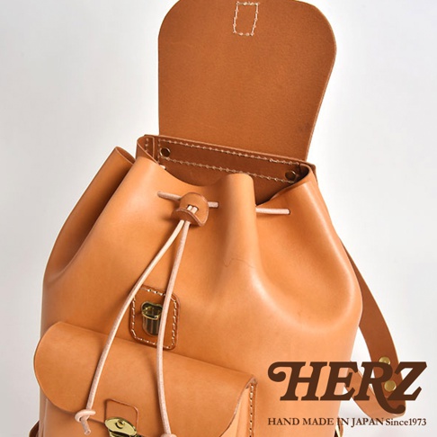 淺棕色皮革後背包，束口設計，正面有翻蓋口袋，黃銅色金屬扣環，簡約經典風格。