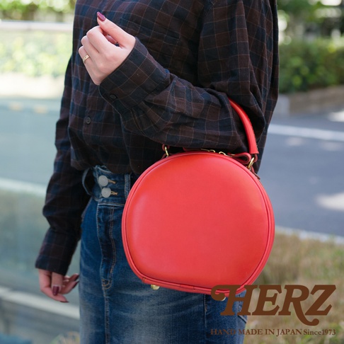 一位女性穿著格紋襯衫和牛仔褲，手裡提著一個紅色的圓形皮革側背包，包包上有 HERZ 的品牌標誌，標誌下方寫著 HAND MADE IN JAPAN Since1973