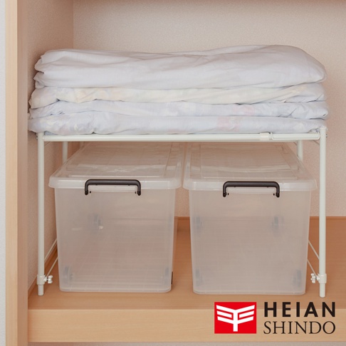 白色金屬雙向可伸縮置物整理架，架上擺放著疊好的白色棉被，下方放置兩個透明塑膠收納箱，每個收納箱都有黑色提把。