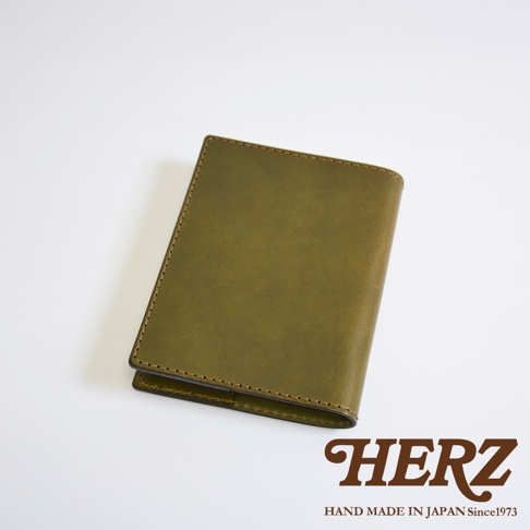 這是一款日本職人品牌 HERZ 手作的橄欖綠色皮革書套，外觀呈現簡約質樸的厚實質感，表面有細緻皮革紋理，邊緣以精細車縫線加固，右下角印有 HERZ HAND MADE IN JAPAN Since1973 品牌標誌。