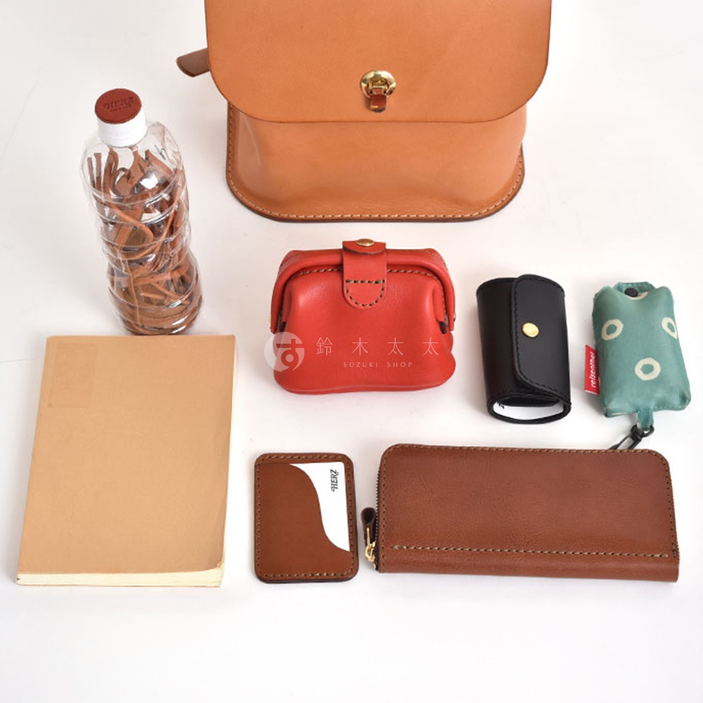 淺棕色皮革翻蓋迷你後背包，搭配金色鎖扣，旁邊擺放著一瓶水、紅色皮革零錢包、黑色鑰匙包、綠色印花收納袋、筆記本、棕色名片夾和長款棕色皮夾。