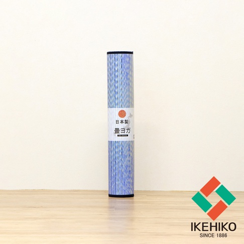 捲筒狀的藺草瑜珈墊，表面呈現藍白交織的細緻紋理，帶有日文標籤，置於淺木色地板上，背景為米白色牆面。