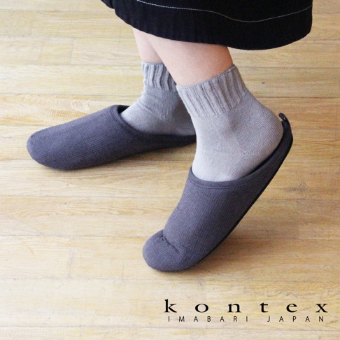 一雙深灰色、素面的 MOKU 棉麻室內拖鞋，鞋面帶有細緻的織紋，由穿著灰色短襪的雙腳穿著，置於淺木紋地板上。