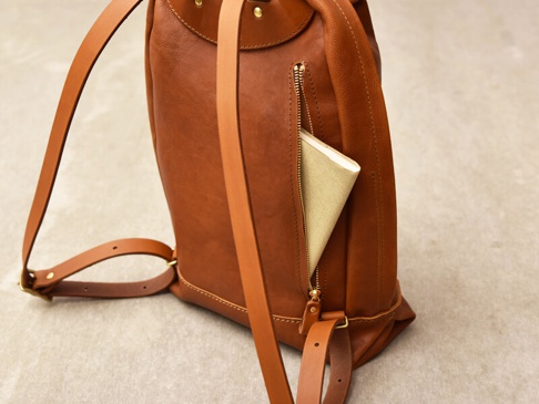 職人手作皮革後背包，色澤為溫暖的棕色，材質為真皮。包體帶有簡約的褶皺設計，側邊有一個拉鍊口袋，內有放置一本淺黃色筆記本。包款配有可調整的皮革肩帶和金屬扣環。